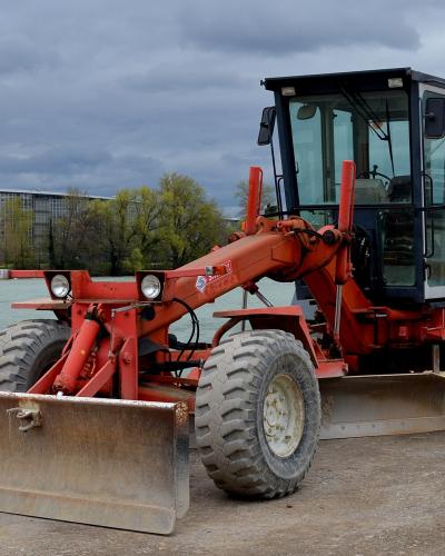A red O&K motor grader parked on a dirt construction site next to a river, with a large modern building under construction visible in the background across the water.