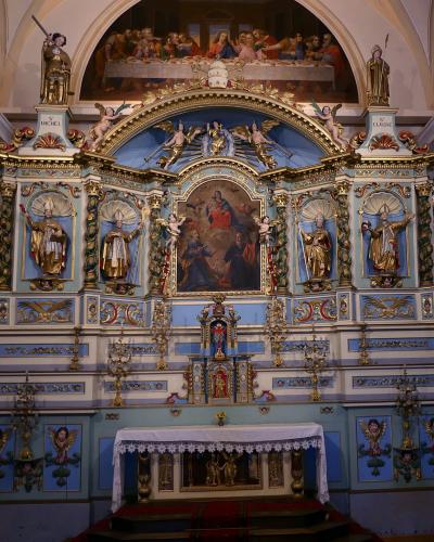 An ornate, light-blue and gold altarpiece (retable) inside a church, featuring statues of saints, a central painting of the Last Supper, and inscriptions reading "S. MICHEL" and "S. CLAUDE".
