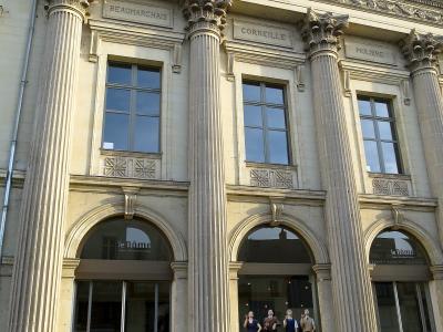 The image shows the neoclassical facade of a theater building featuring large Corinthian columns, inscriptions of playwrights (Beaumarchais, Corneille, Molière), and a blue bull sculpture in front, with signage reading "le Dôme" above the arched e