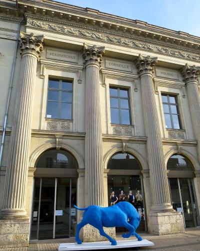 The image shows the neoclassical facade of a theater building featuring large Corinthian columns, inscriptions of playwrights (Beaumarchais, Corneille, Molière), and a blue bull sculpture in front, with signage reading "le Dôme" above the arched e