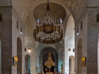 The interior of a large stone cathedral featuring massive piers, rounded arches, and a prominent central dome, with two large ornate chandeliers hanging from the ceiling.