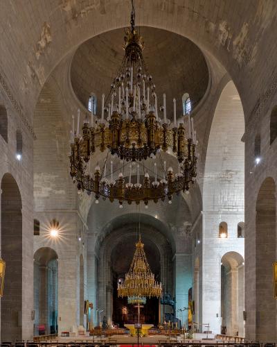 The interior of a large stone cathedral featuring massive piers, rounded arches, and a prominent central dome, with two large ornate chandeliers hanging from the ceiling.