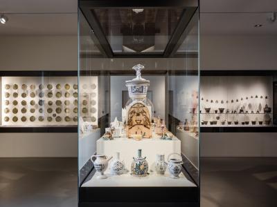The image shows a modern museum interior with a central glass display case containing ceramic vessels and a large ornate jar, flanked by wall-mounted display niches holding rows of coins and pottery, under track lighting.