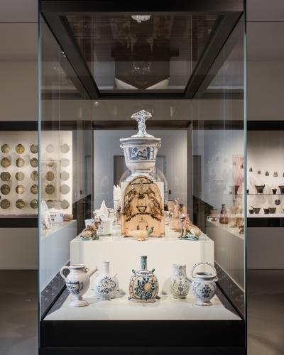 The image shows a modern museum interior with a central glass display case containing ceramic vessels and a large ornate jar, flanked by wall-mounted display niches holding rows of coins and pottery, under track lighting.