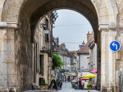 A stone archway gateway leading onto a cobblestone street in a historic French town, with buildings, market umbrellas, and a blue directional traffic sign visible through the arch.