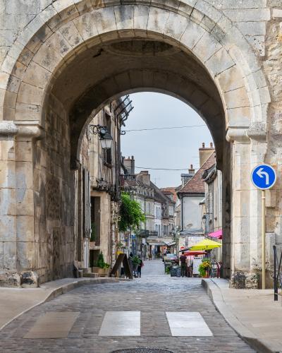 A stone archway gateway leading onto a cobblestone street in a historic French town, with buildings, market umbrellas, and a blue directional traffic sign visible through the arch.