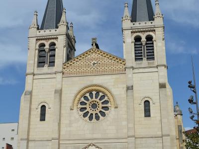 A low-angle photograph of a large, light-colored stone church with two symmetrical bell towers topped by dark spires and crosses, featuring a prominent rose window and an ornate central portal.