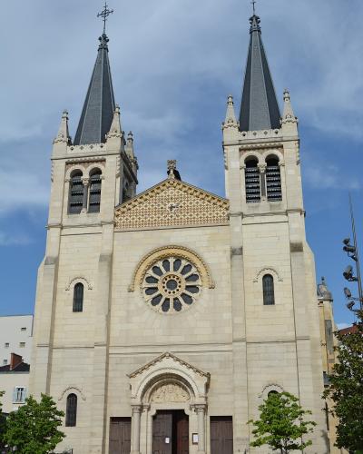 A low-angle photograph of a large, light-colored stone church with two symmetrical bell towers topped by dark spires and crosses, featuring a prominent rose window and an ornate central portal.