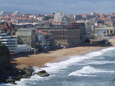 A high-angle view of a sandy beach curving along the coast, backed by a dense urban skyline of multi-story buildings with terracotta roofs and prominent hotels, including a large red-and-white facade building near the center; waves break on the sh