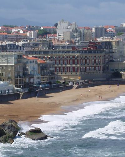 A high-angle view of a sandy beach curving along the coast, backed by a dense urban skyline of multi-story buildings with terracotta roofs and prominent hotels, including a large red-and-white facade building near the center; waves break on the sh