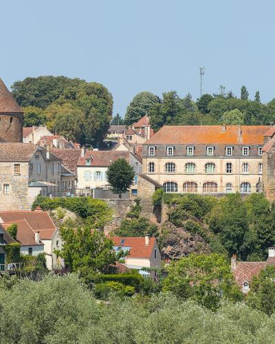 A panoramic view of the Château de Semur-en-Auxois, featuring its distinctive round towers with conical roofs and stone walls, situated on a rocky outcrop above the town, viewed from a distance across greenery.