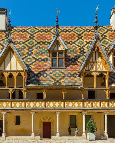 A close-up architectural detail of a gable featuring a carved cherub holding a shield, topped with a sunburst motif, set against a background of colorful glazed tiles in yellow, green, and black.