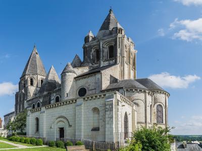 The image shows the exterior of a large Romanesque stone church with multiple conical slate roofs, towers, and an apse, set against a blue sky with green landscaping in the foreground.