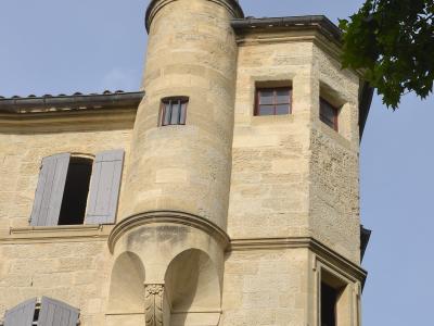 A low-angle view of a historic stone building in Uzès, France, featuring a prominent cylindrical corner turret with a domed roof, arched corbels, and shuttered windows under a clear blue sky.