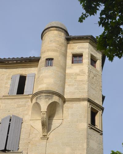 A low-angle view of a historic stone building in Uzès, France, featuring a prominent cylindrical corner turret with a domed roof, arched corbels, and shuttered windows under a clear blue sky.