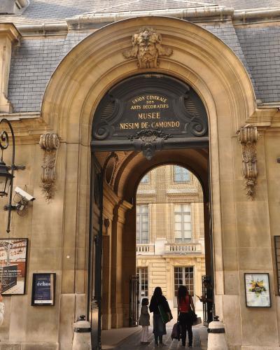 A low-angle street view showing a classic Parisian Haussmann-style building on the left, a blue circular street sign reading "MUSÉE NISSIM DE CAMONDO" with the address "63 rue de Monceau", and a black Morris column advertising "SPECTACLES" on the