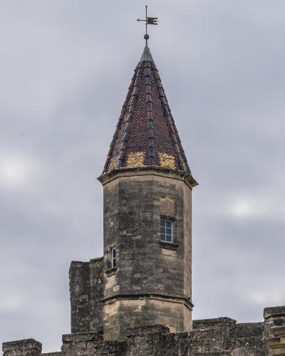 A stone tower with a conical tiled roof and a weather vane, part of a larger fortified stone structure.