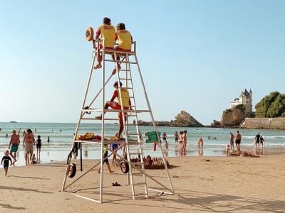 A tall white lifeguard tower stands on a sandy beach with several people in yellow and red uniforms sitting or climbing it; the background shows the ocean, swimmers, and a distinctive castle-like building on a rocky headland to the right.