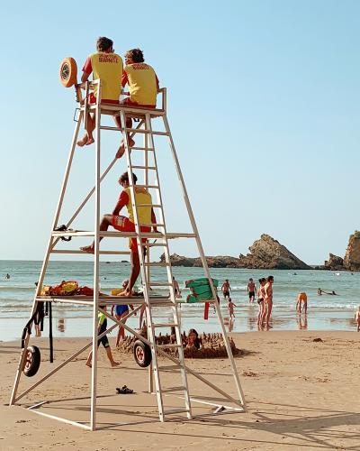 A tall white lifeguard tower stands on a sandy beach with several people in yellow and red uniforms sitting or climbing it; the background shows the ocean, swimmers, and a distinctive castle-like building on a rocky headland to the right.