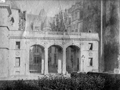 A black-and-white photograph showing a stone arcade with three arches and a balustrade above, part of a larger castle complex with towers and turrets in the background.