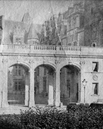 A black-and-white photograph showing a stone arcade with three arches and a balustrade above, part of a larger castle complex with towers and turrets in the background.