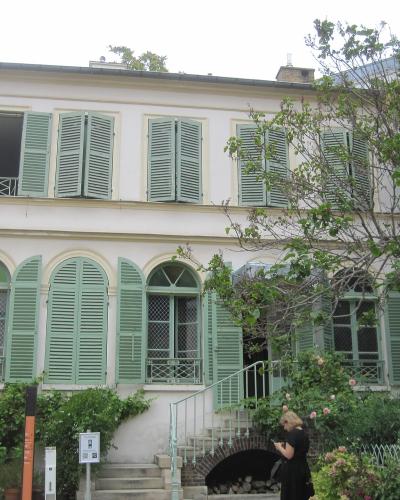 A cream-colored building with brown shutters and a white canopy tent in the foreground, part of a courtyard or garden area with adjacent stone walls and a taller white building in the background.