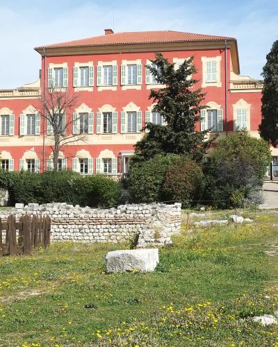 A large, salmon-pink villa with white shutters and a terracotta roof, situated behind ancient stone ruins and a grassy field with yellow flowers under a blue sky.