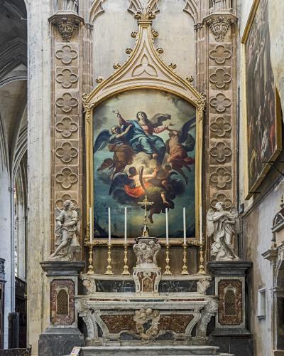 The image shows an ornate Baroque-style altar with a large painting of the Assumption of the Virgin Mary, flanked by two angel statues and set within a stone architectural niche featuring quatrefoil motifs.