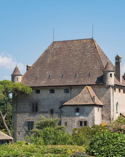 A large medieval stone castle with a steep tiled roof, corner turrets, and a smaller attached building, viewed from a garden with greenery in the foreground and a lake and distant hills in the background under a blue sky.