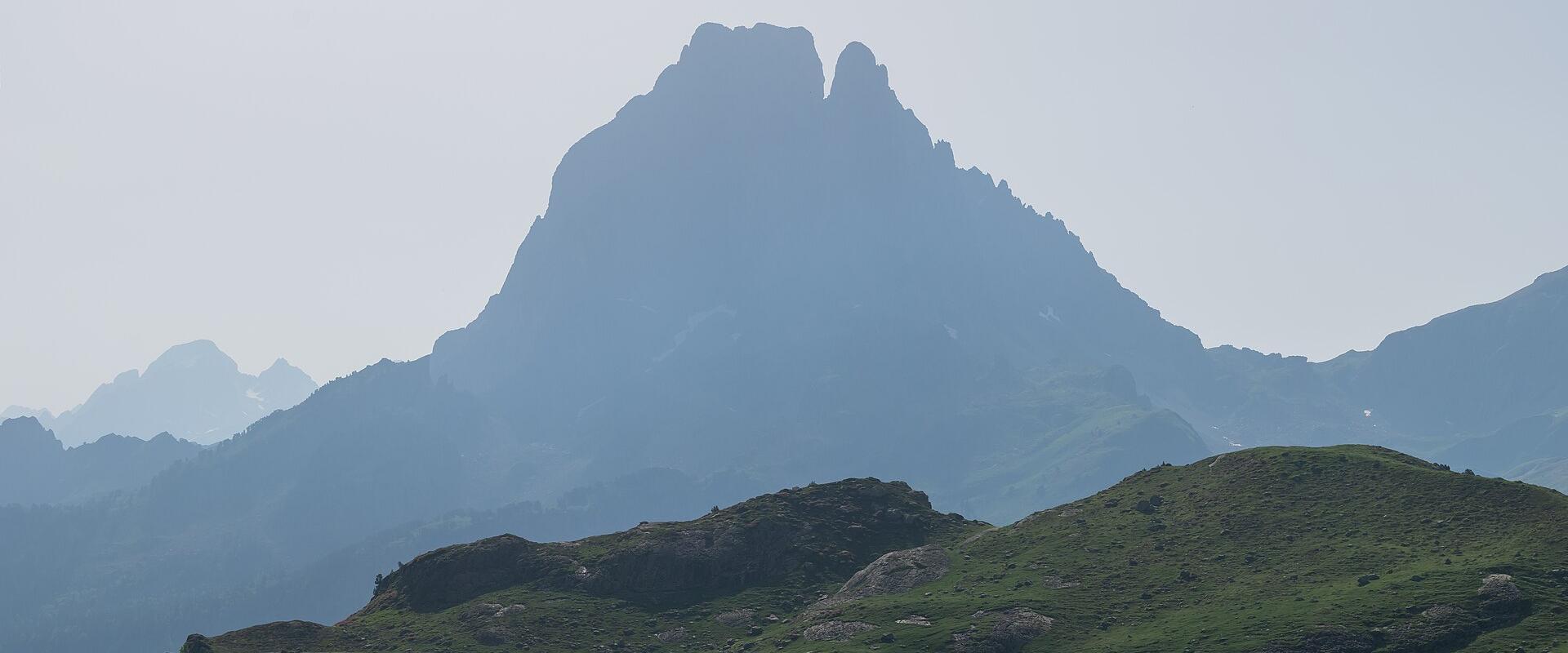 1. A mountain with a distinctive double-peak silhouette rises above a green alpine meadow and a lake in the foreground, under a hazy sky.