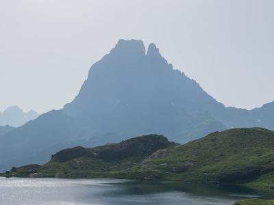 1. A mountain with a distinctive double-peak silhouette rises above a green alpine meadow and a lake in the foreground, under a hazy sky.