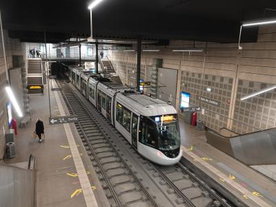 A modern metro station platform with a white tram/train stopped at the platform, featuring signage reading "Palais de Justice" and "Sortie," along with passengers, stairs, escalators, and overhead lighting.