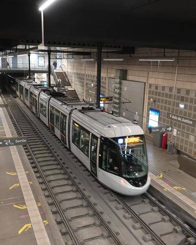 A modern metro station platform with a white tram/train stopped at the platform, featuring signage reading "Palais de Justice" and "Sortie," along with passengers, stairs, escalators, and overhead lighting.