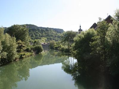 A river flowing through a town with lush green trees on the banks, a church steeple visible on the right, and a rocky cliff face in the background.