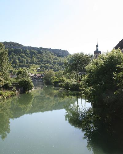 A river flowing through a town with lush green trees on the banks, a church steeple visible on the right, and a rocky cliff face in the background.