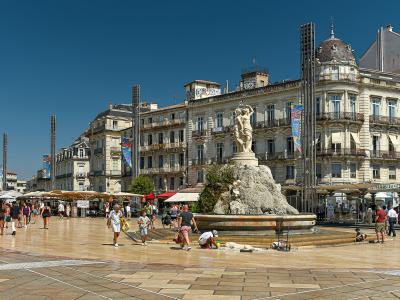 The image shows a large, sunlit urban plaza with a central fountain featuring a statue of a figure on a rocky base, surrounded by multi-story classical-style buildings, outdoor café seating, and tall metal light poles under a clear blue sky.