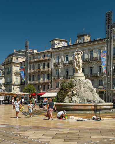The image shows a large, sunlit urban plaza with a central fountain featuring a statue of a figure on a rocky base, surrounded by multi-story classical-style buildings, outdoor café seating, and tall metal light poles under a clear blue sky.