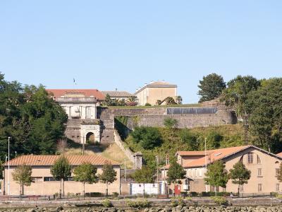 A stone fortress with a prominent arched gateway and terraced ramparts sits atop a grassy hill, overlooking a waterfront promenade with buildings featuring terracotta roofs and a French flag flying.