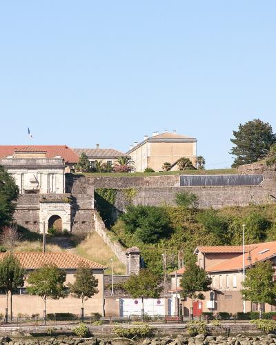 A stone fortress with a prominent arched gateway and terraced ramparts sits atop a grassy hill, overlooking a waterfront promenade with buildings featuring terracotta roofs and a French flag flying.
