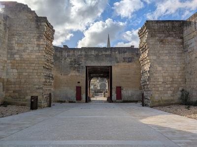 A stone gateway with red doors, flanked by ruined walls, leading into a courtyard where a cathedral spire is visible in the distance under a cloudy sky.