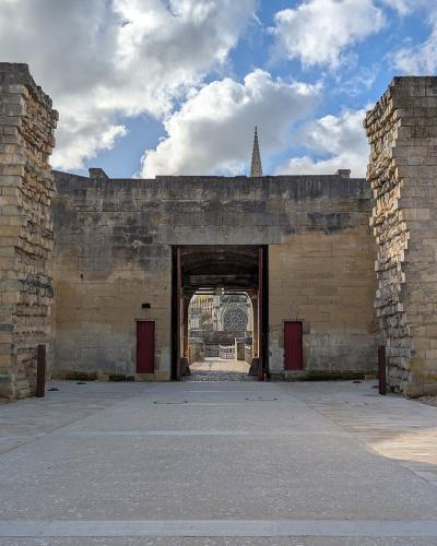 A stone gateway with red doors, flanked by ruined walls, leading into a courtyard where a cathedral spire is visible in the distance under a cloudy sky.