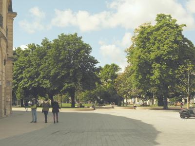 A wide, paved public square or park area with large green trees, people walking, and a building facade on the left under a partly cloudy sky.