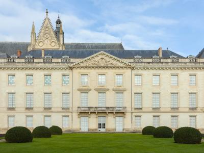A symmetrical, light-colored stone building with a central pediment, multiple rows of tall windows, and manicured round hedges on a lawn in the foreground, with a Gothic-style cathedral spire visible behind it.