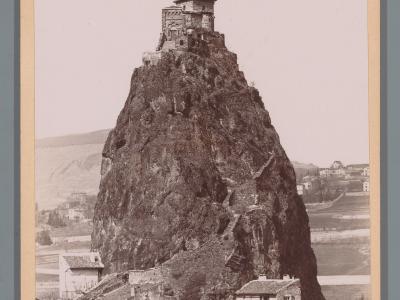 A vintage sepia-toned photograph showing a small stone chapel with a bell tower perched atop a steep, isolated volcanic rock pinnacle, with houses at its base and rolling hills in the background; no visible text or signage within the image itself.