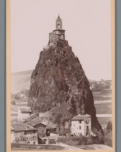 A vintage sepia-toned photograph showing a small stone chapel with a bell tower perched atop a steep, isolated volcanic rock pinnacle, with houses at its base and rolling hills in the background; no visible text or signage within the image itself.
