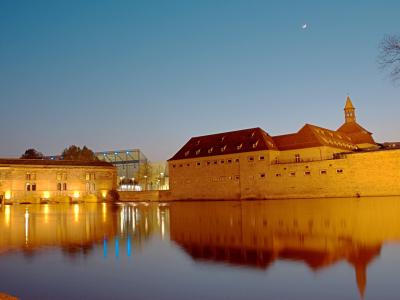 A twilight view of the historic fortifications and buildings along the Ill River in Strasbourg, featuring the illuminated stone walls of the former barracks (now part of the ENA campus) and the modern glass structure of the MAMCS in the background