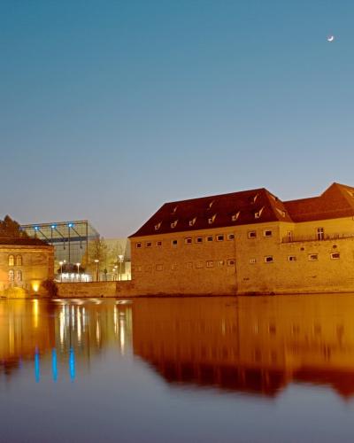 A twilight view of the historic fortifications and buildings along the Ill River in Strasbourg, featuring the illuminated stone walls of the former barracks (now part of the ENA campus) and the modern glass structure of the MAMCS in the background