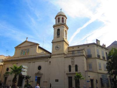 The image shows a light-colored stone church with a prominent bell tower topped by a dome and cross, a clock face on the tower, a triangular pediment with a mosaic above the entrance, and street signs in French (including "LE CHAPEAU" and "PARCOUR