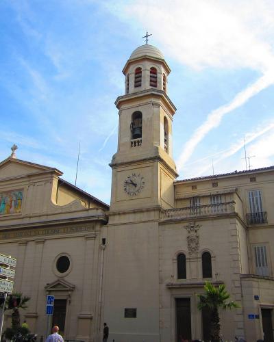 The image shows a light-colored stone church with a prominent bell tower topped by a dome and cross, a clock face on the tower, a triangular pediment with a mosaic above the entrance, and street signs in French (including "LE CHAPEAU" and "PARCOUR