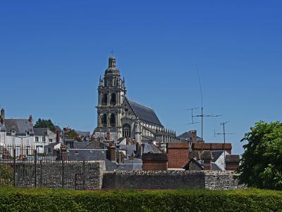 A large stone cathedral with a prominent central tower and dome, viewed from a distance over rooftops and a stone wall under a clear blue sky.